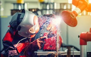 Soldador con careta trabajando junto a un equipo robotizado en una instalación industrial
